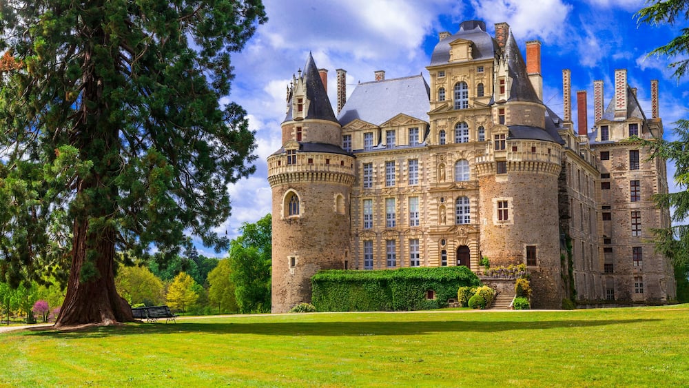 Un vaste château historique, doté de nombreuses tours et tourelles, entouré d’une végétation luxuriante et d’une pelouse bien entretenue.