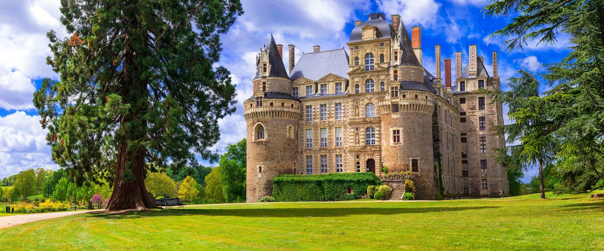 A large, historic castle with multiple towers and turrets, surrounded by lush greenery and a well-maintained lawn.