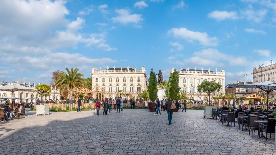 A historic plaza with ornate buildings, cobblestone pavement, and outdoor seating areas.