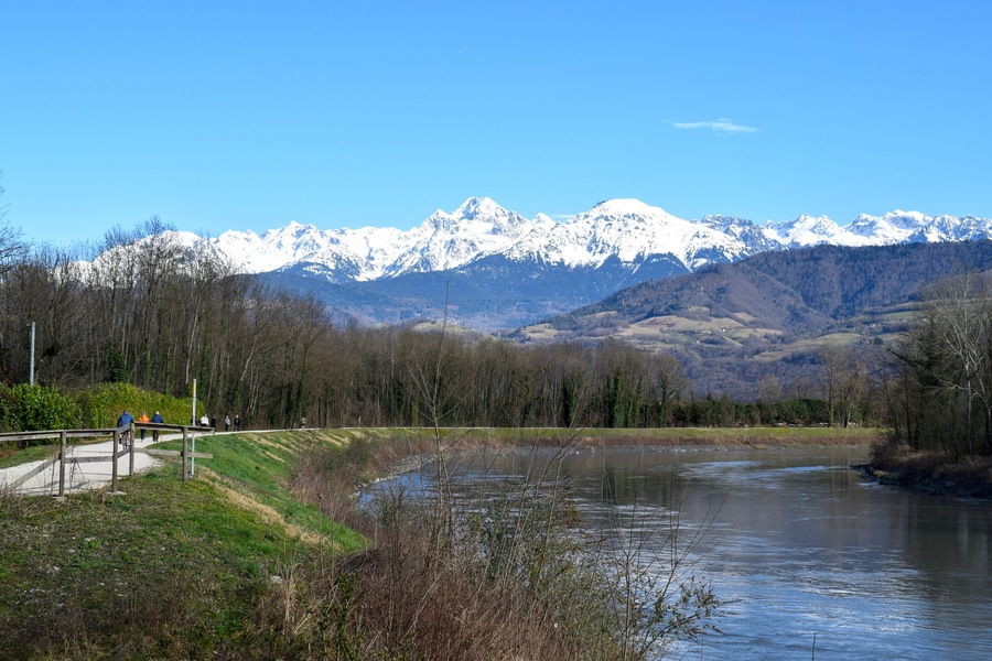 People strolling along the Isere River in Saint-Martin-d'Heres, with stunning snow-capped Alps in the background. A peaceful mountain landscape.