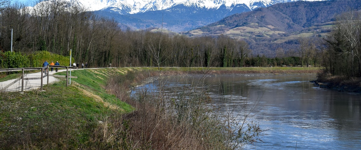 People strolling along the Isere River in Saint-Martin-d'Heres, with stunning snow-capped Alps in the background. A peaceful mountain landscape.