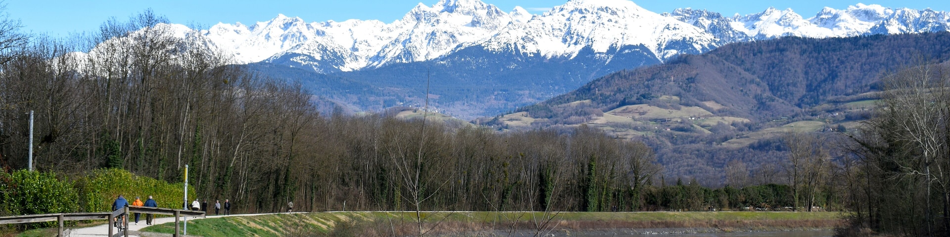 People strolling along the Isere River in Saint-Martin-d'Heres, with stunning snow-capped Alps in the background. A peaceful mountain landscape.