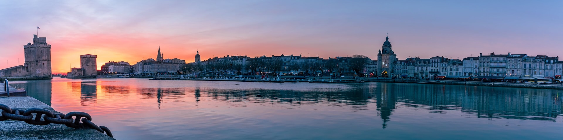 Ein Stadtpanorama bei Sonnenuntergang, wobei ein ruhiger Fluss die Gebäude spiegelt, ein markanter Turm und eine Brücke zu sehen sind.
