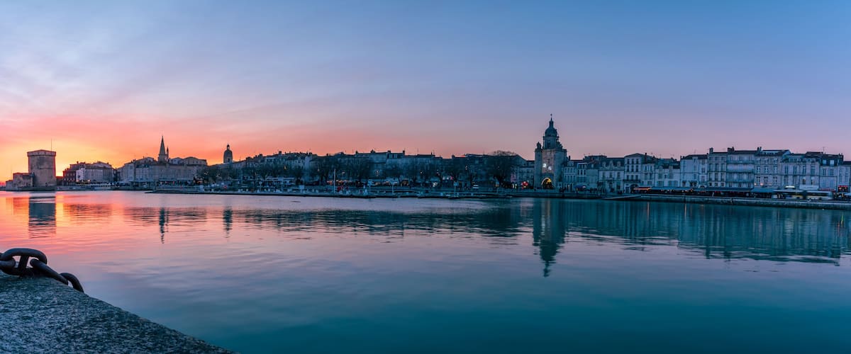 A city skyline at sunset with a calm river reflecting the buildings, a prominent tower, and a bridge.