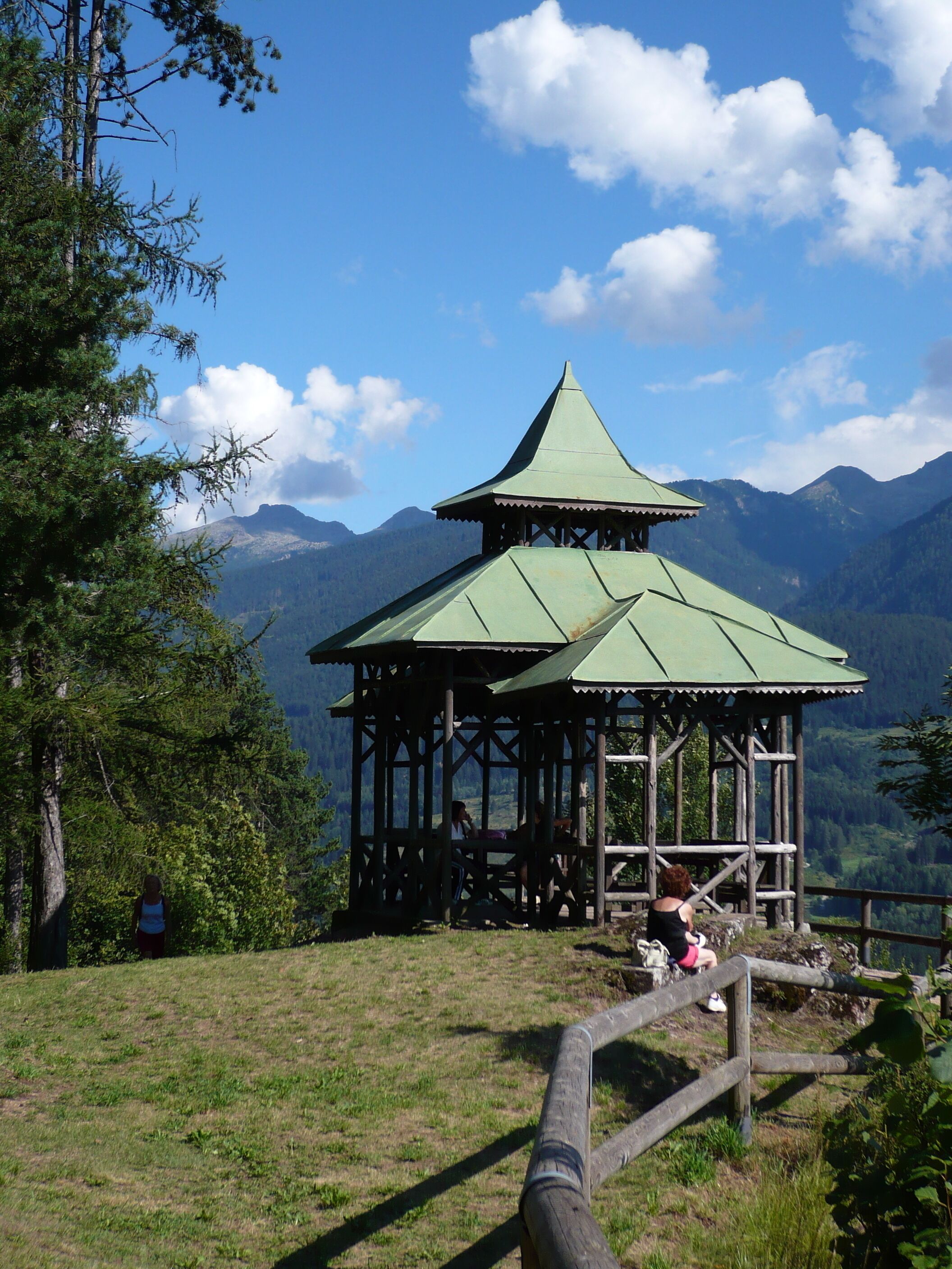 Um gazebo de madeira tradicional com telhado verde, rodeado por vegetação exuberante e montanhas ao fundo.