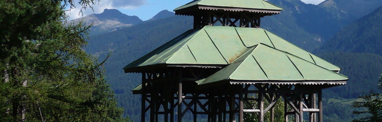 Um gazebo de madeira tradicional com telhado verde, rodeado por vegetação exuberante e montanhas ao fundo.
