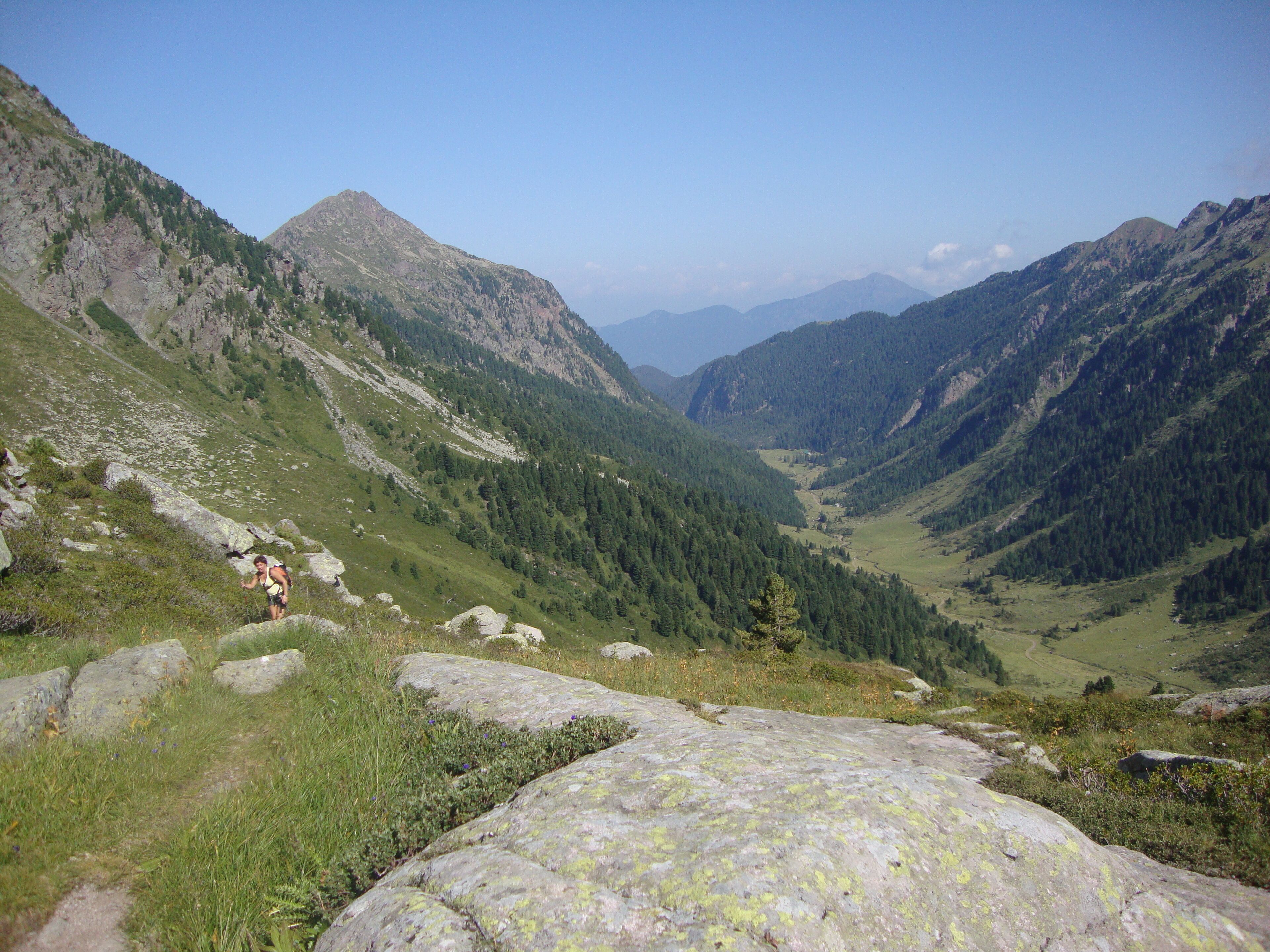 A hiker on a rocky path in a mountainous valley with dense forests and distant peaks.