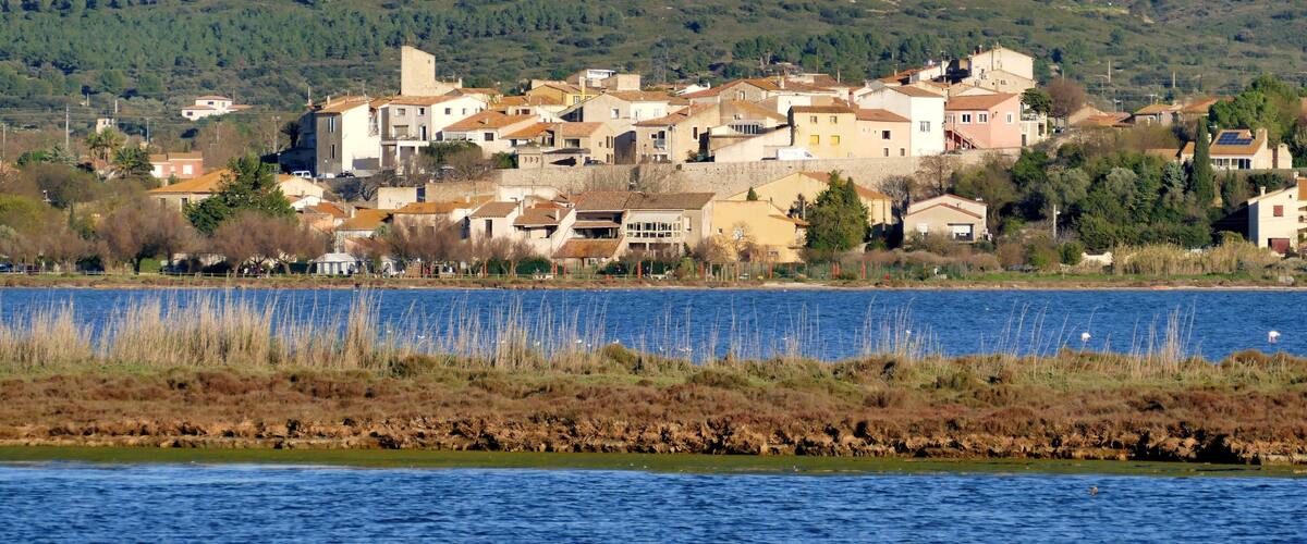 A coastal town with buildings along the shoreline, a body of water in the foreground, and a hilly landscape in the background.