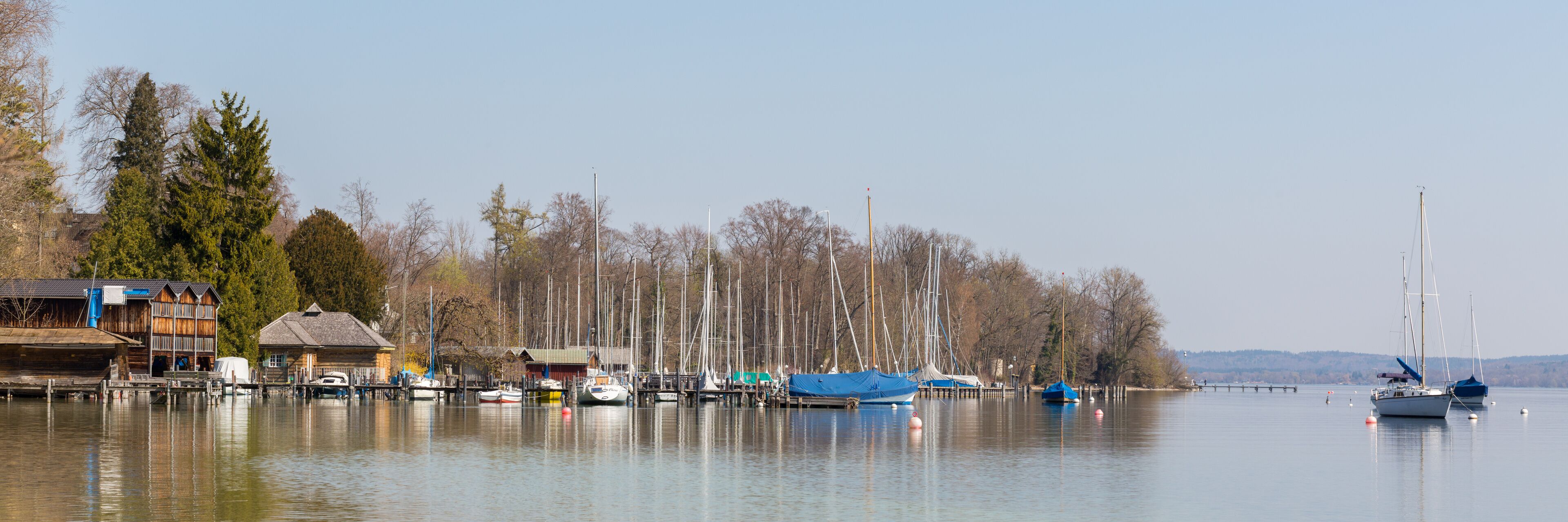 A calm lake with boats docked along the shore, surrounded by trees and buildings.