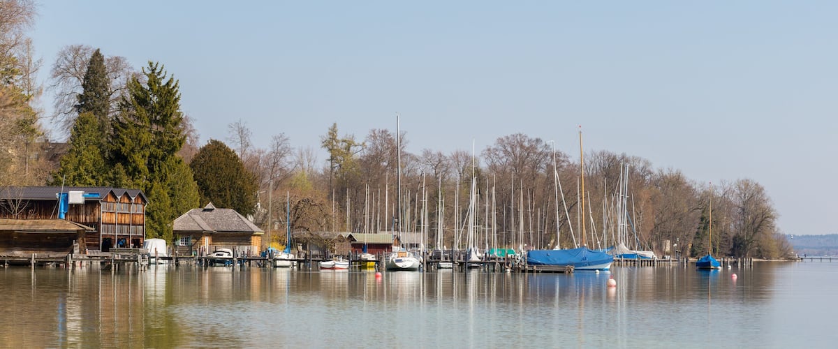 A calm lake with boats docked along the shore, surrounded by trees and buildings.