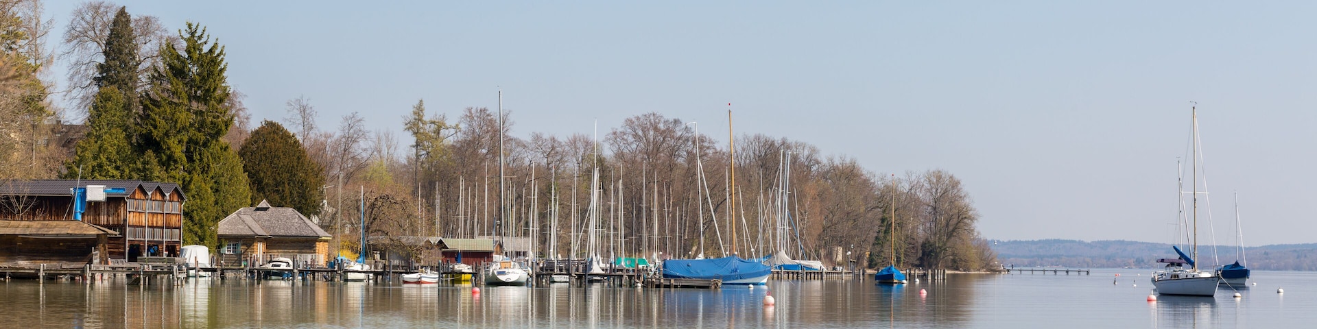 A calm lake with boats docked along the shore, surrounded by trees and buildings.