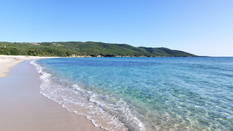 A beach with clear blue water, sandy shore, and green hills in the background.