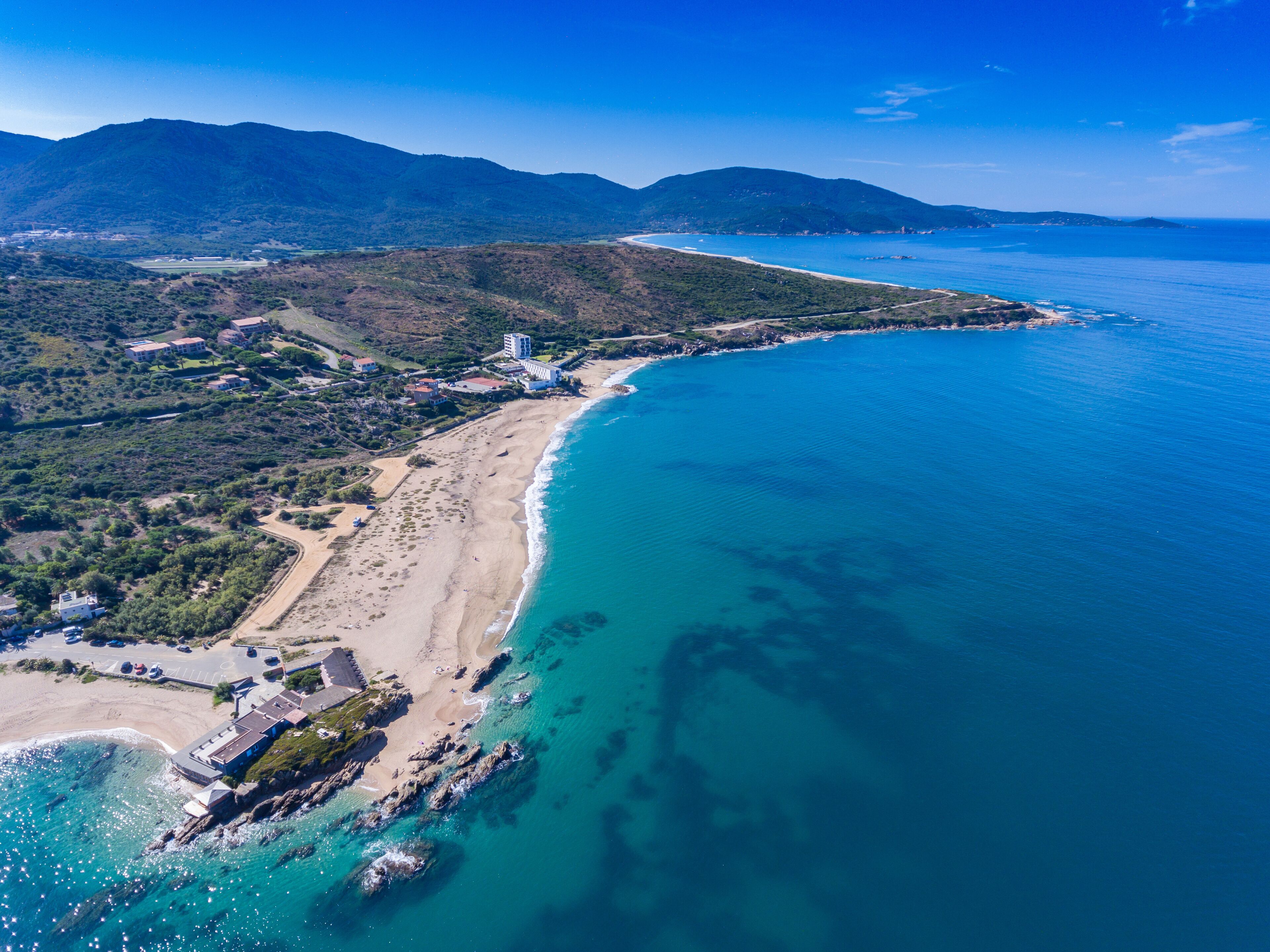 A coastal landscape with a sandy beach, clear blue waters, and a mountainous backdrop.