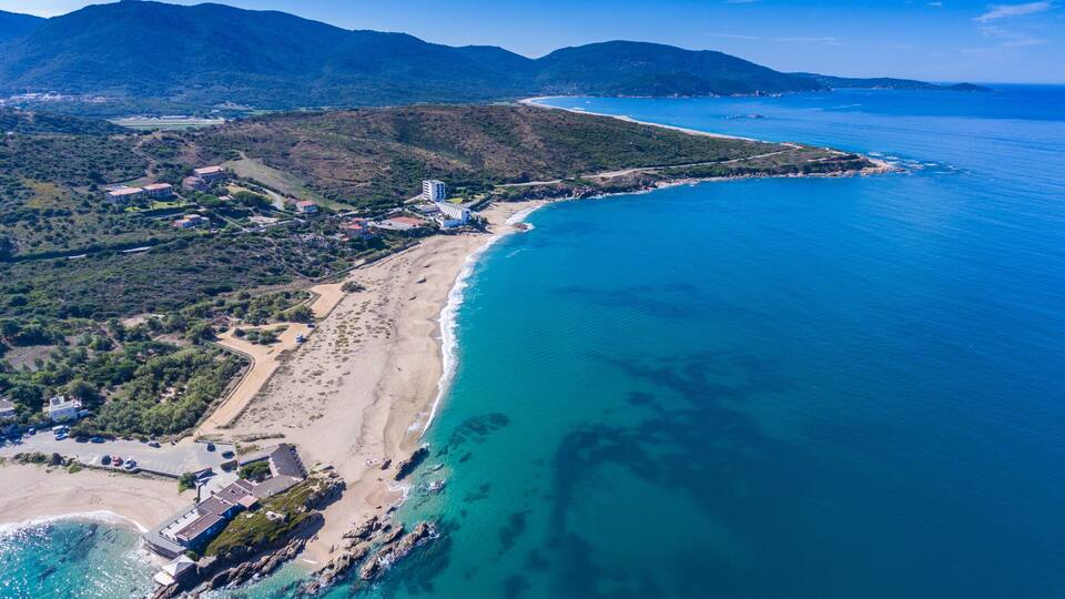 A coastal landscape with a sandy beach, clear blue waters, and a mountainous backdrop.