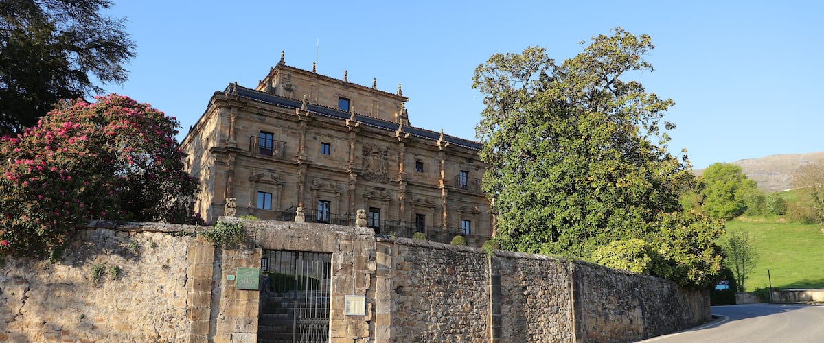 A historic stone building with a tiled roof, surrounded by lush greenery and a stone wall.