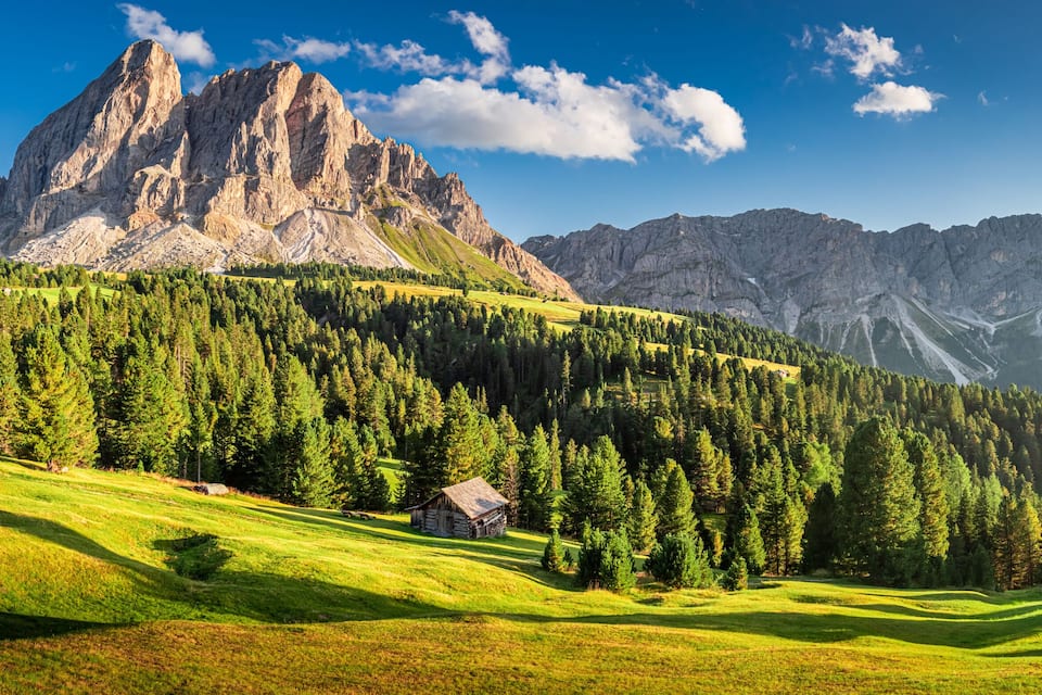 A mountainous landscape with a prominent rocky peak, dense forested slopes, and a small cabin.