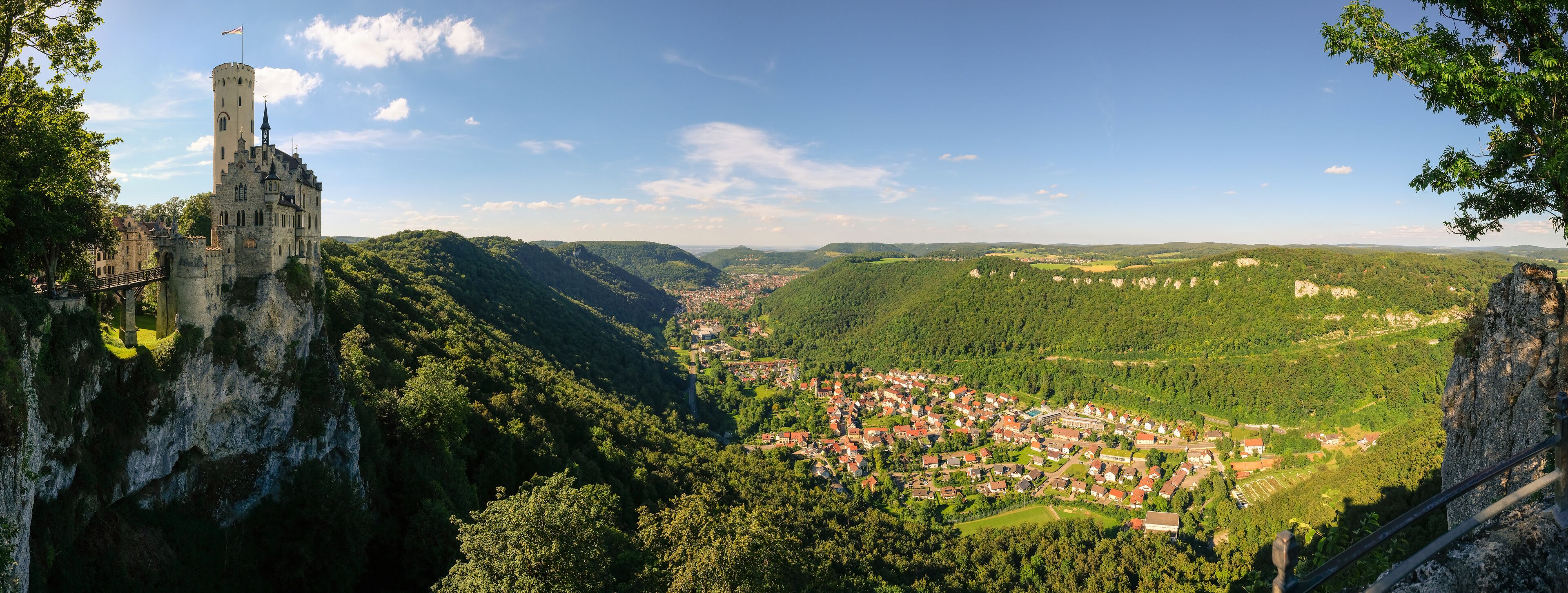 A castle on a cliff overlooking a village surrounded by forested hills.