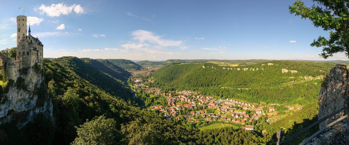 A castle on a cliff overlooking a village surrounded by forested hills.