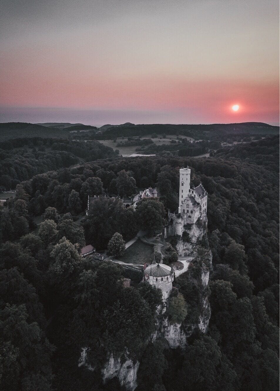 Um castelo rodeado por uma floresta densa, com um pôr do sol ao fundo.