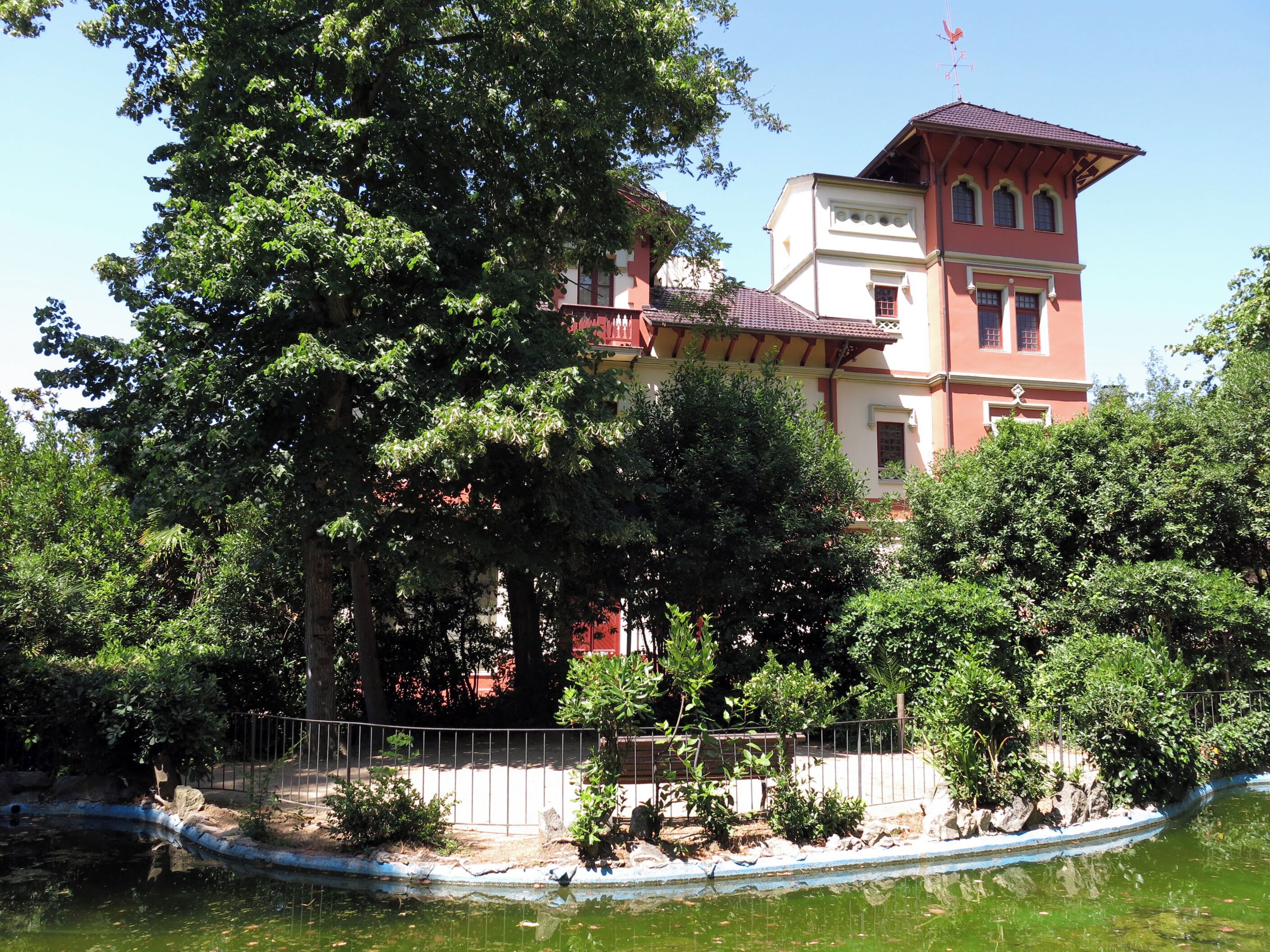 A large tree in front of a building with a red roof and white walls.