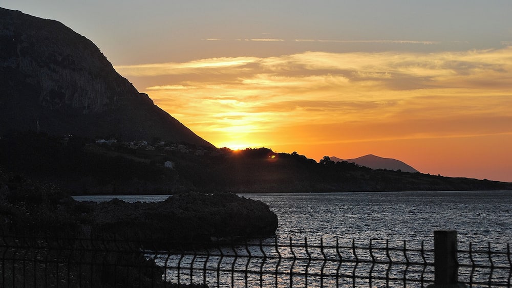 A sunset over a coastal landscape with a mountain in the background and a fence in the foreground.