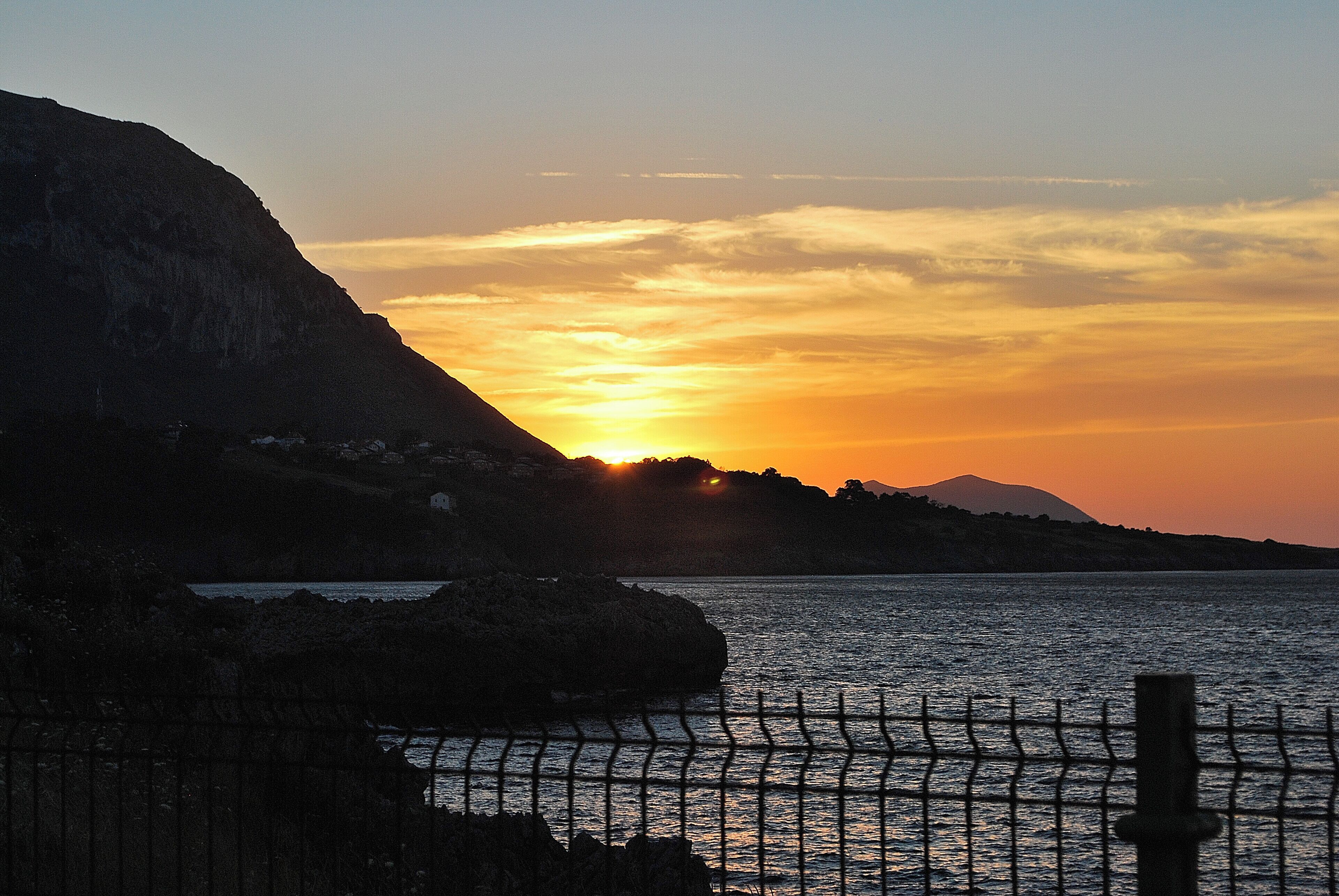 A sunset over a coastal landscape with a mountain in the background and a fence in the foreground.