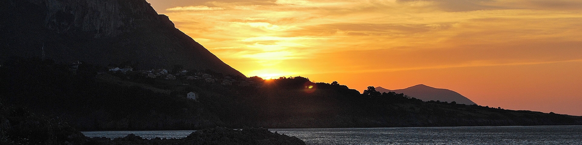 A sunset over a coastal landscape with a mountain in the background and a fence in the foreground.