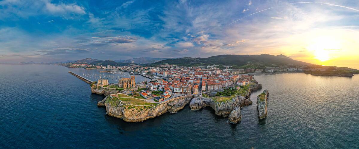 A coastal cityscape with a prominent cathedral, a pier, and a rocky peninsula.