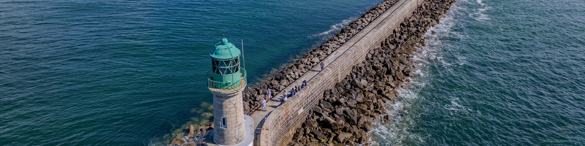 A lighthouse with a green top, a long stone breakwater, and calm blue waters.