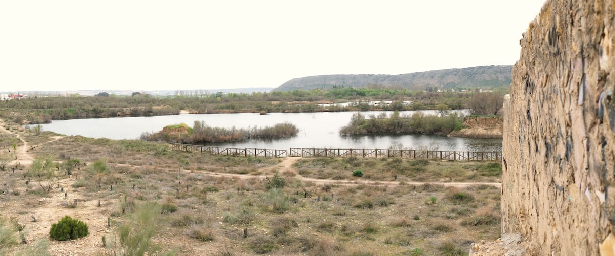 A river with a wooden bridge, surrounded by sparse vegetation and distant hills.