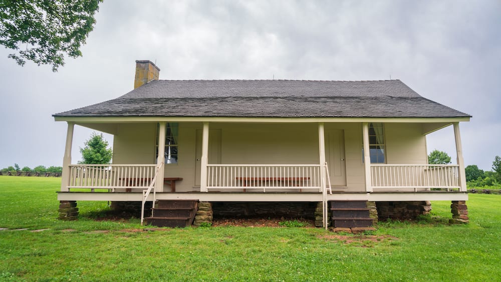 A single-story house with a porch, stone foundation, and a grassy field in the background.