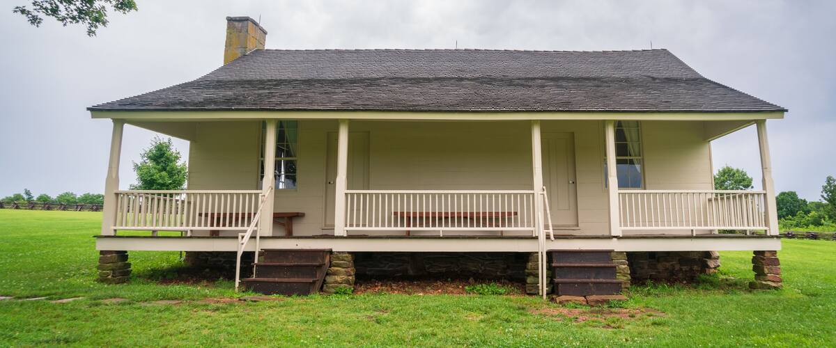 A single-story house with a porch, stone foundation, and a grassy field in the background.