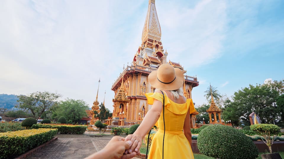A woman in a yellow dress and a straw hat holding hands with a man, standing in front of a temple with a tall spire.
