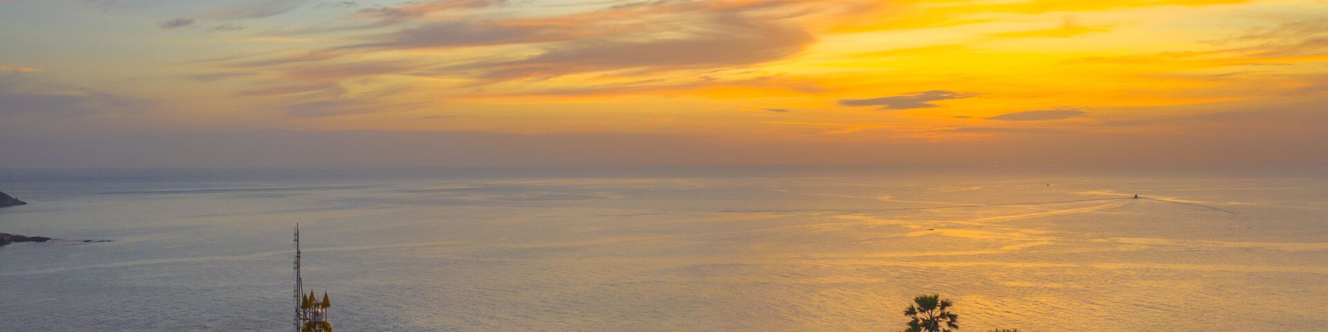 Um pôr do sol costeiro com um farol, palmeiras e uma praia cheia de pessoas.