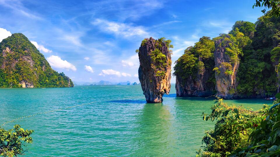 A clear turquoise sea with a prominent rock formation in the center, surrounded by lush green hills and a partly cloudy sky.