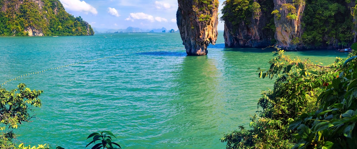 A clear turquoise sea with a prominent rock formation in the center, surrounded by lush green hills and a partly cloudy sky.