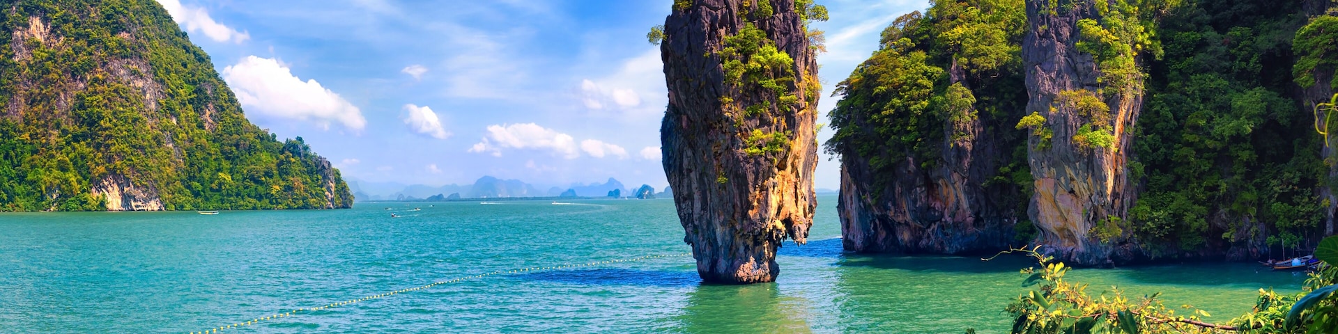 A clear turquoise sea with a prominent rock formation in the center, surrounded by lush green hills and a partly cloudy sky.
