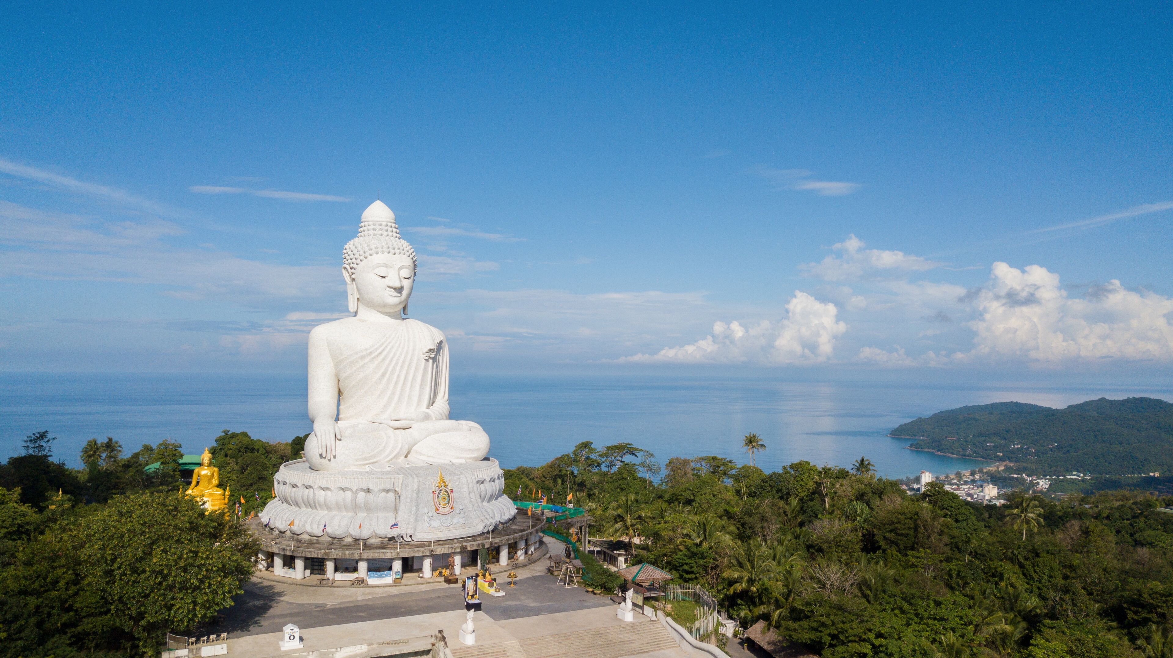A large white Buddha statue surrounded by lush greenery and a clear blue sky.