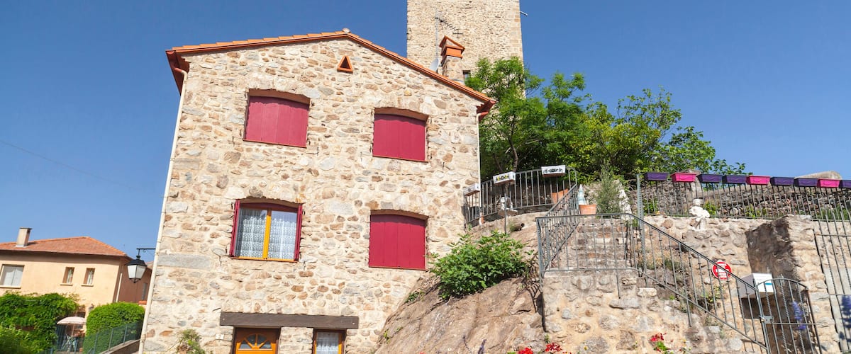 A stone building with red shutters and a yellow door, a stone staircase, and a tower in the background.