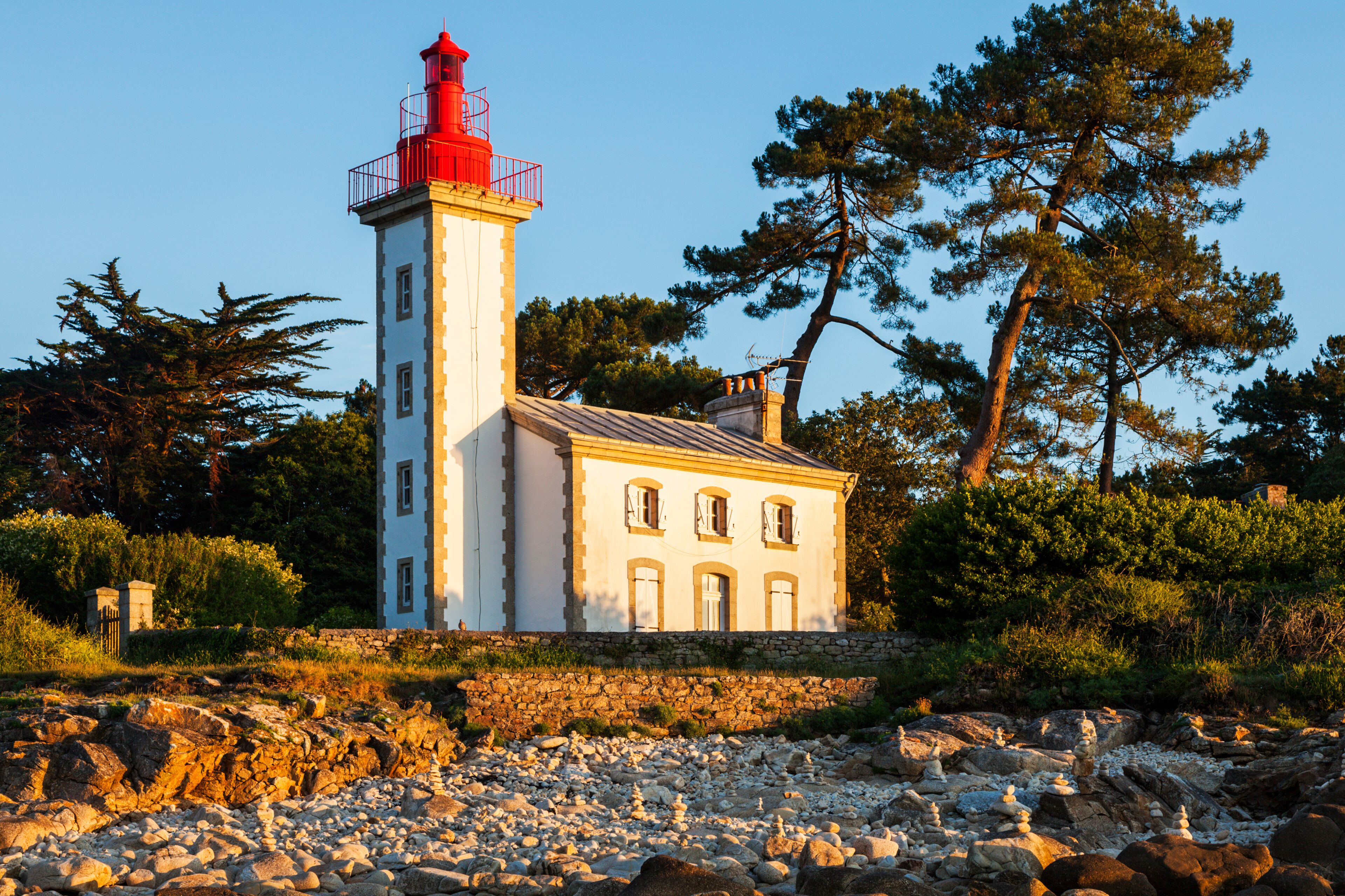 A lighthouse with a red top, a white building with multiple windows, and a rocky shoreline with trees in the background.