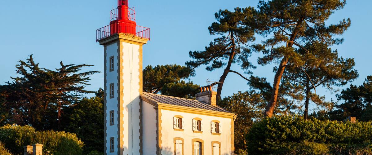 A lighthouse with a red top, a white building with multiple windows, and a rocky shoreline with trees in the background.