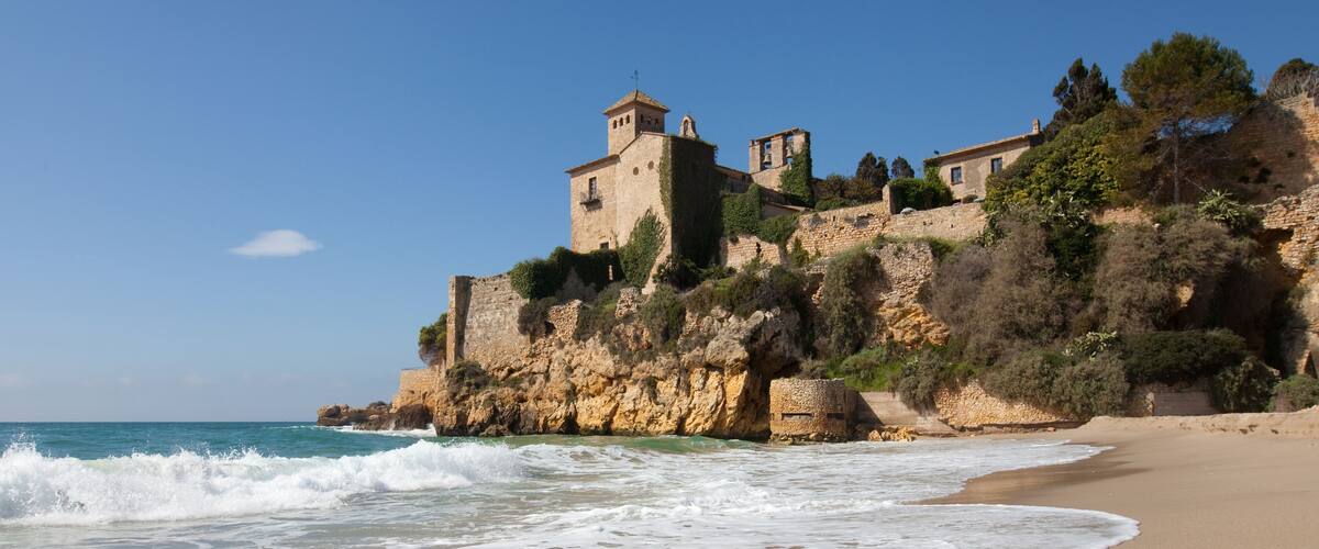 A coastal landscape with a sandy beach, waves, and a historic stone building on a cliff.