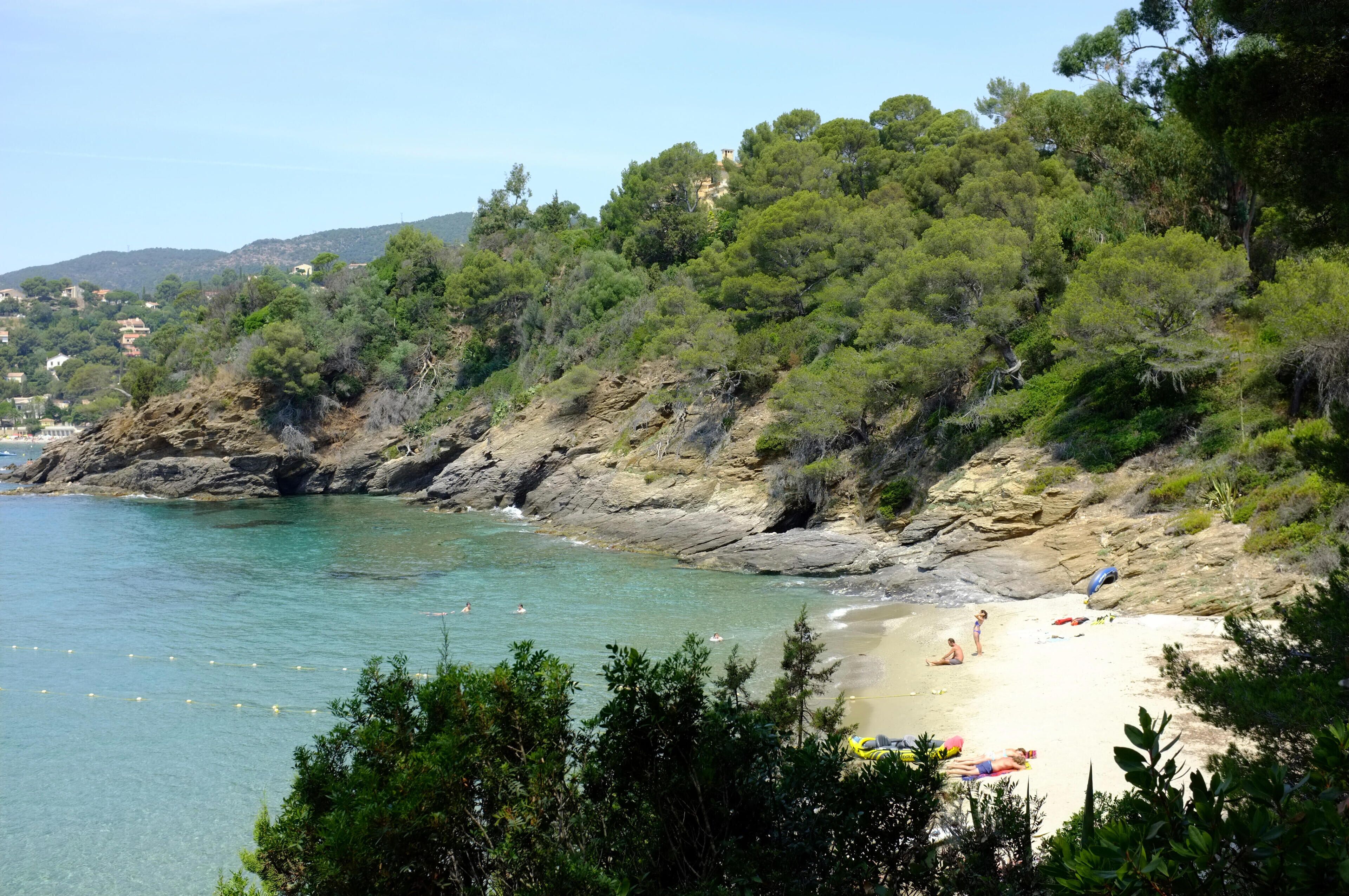 A coastal beach with clear turquoise water, rocky cliffs, and dense green vegetation.
