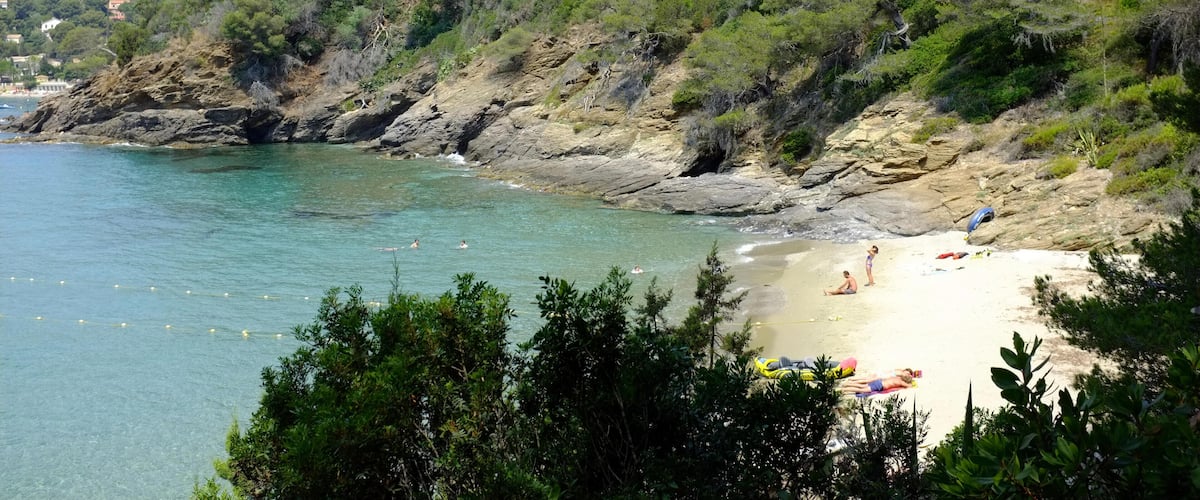 A coastal beach with clear turquoise water, rocky cliffs, and dense green vegetation.