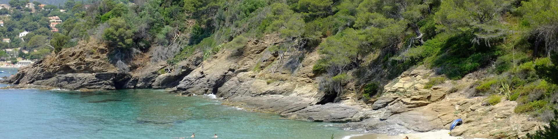 A coastal beach with clear turquoise water, rocky cliffs, and dense green vegetation.