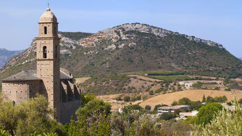 A historic stone church with a bell tower, surrounded by rolling hills and a mountainous backdrop.