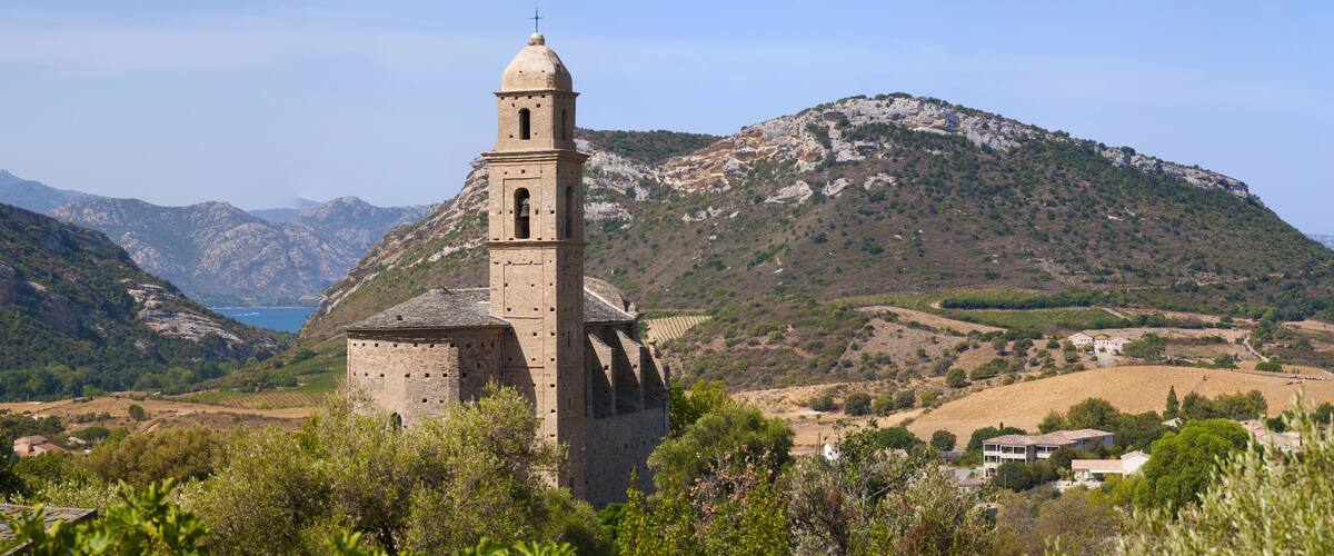 A historic stone church with a bell tower, surrounded by rolling hills and a mountainous backdrop.
