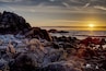 A rocky coastline at sunset with the sun low on the horizon.