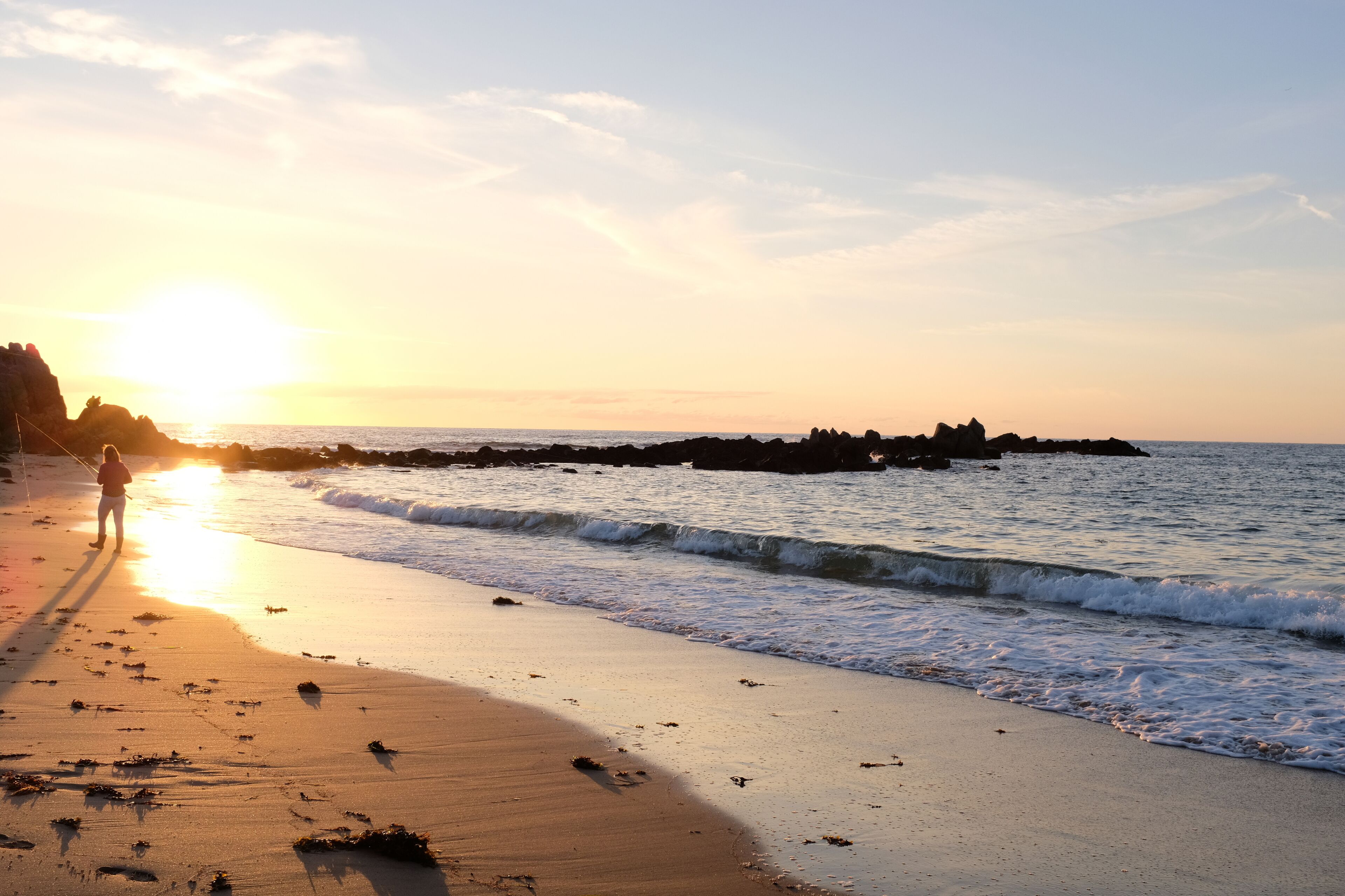 A beach at sunset with waves, a person fishing, and rocky outcrops.