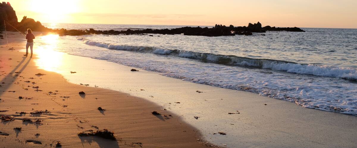 A beach at sunset with waves, a person fishing, and rocky outcrops.